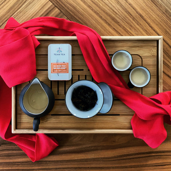 A gaiwan of Da Hong Pao on a wooden tea tray with 2 small cups, a Team Tea slide tin, and a bright red sash draped over.