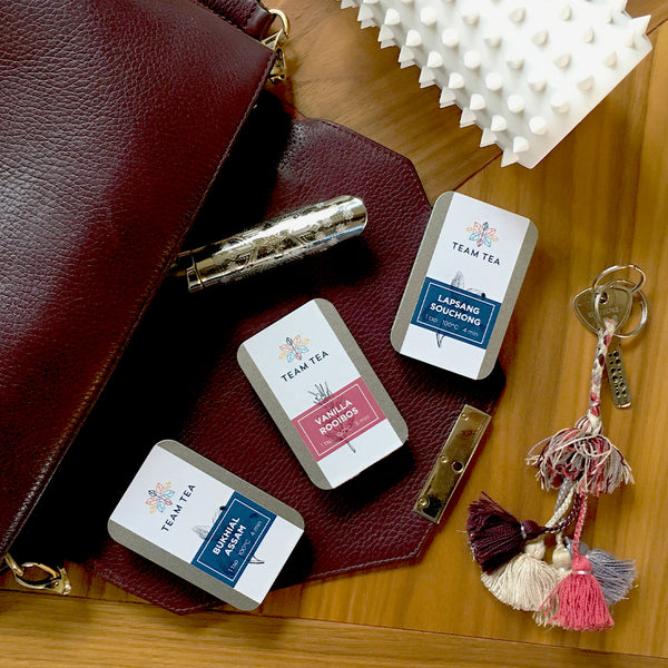 A burgundy handbag on a wooden table, with 3 slide tins of Team Tea spilling out of it. There is also a travel strainer and a house key.