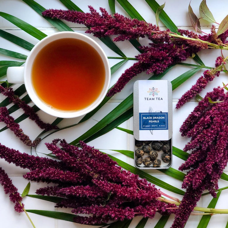 A cup of black tea sits next to an open metal tin of loose-leaf hand rolled black tea pearls. They are surrounded by green palm like foliage and fluffy purple flowers.