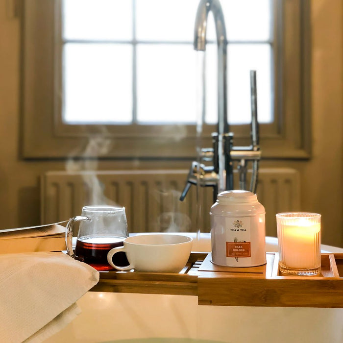 A tray sits a top a bath with a tap in the background. On the tray is a book, a glass teapot of oolong tea, a teacup, a lit candle, and a metal caddy of loose-leaf tea.