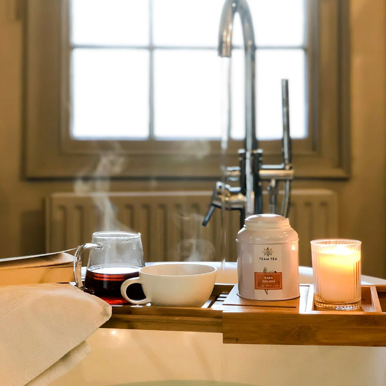 A tray sits a top a bath with a tap in the background. On the tray is a book, a glass teapot of oolong tea, a teacup, a lit candle, and a metal caddy of loose-leaf tea.