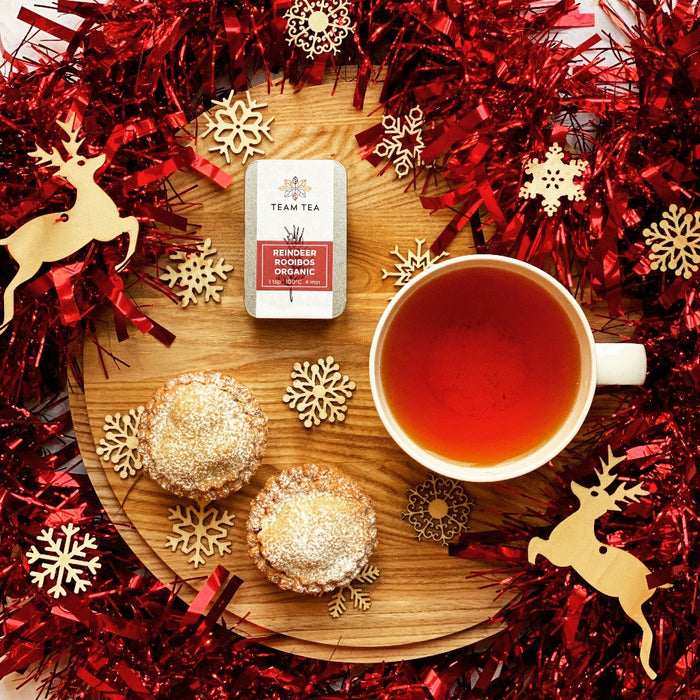 A vibrant cup of Rooibos sits next to 2 mince pies on a wooden board surrounded by Christmas decorations and red tinsel.