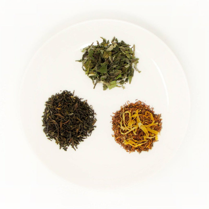 3 small piles of dry leaves of fine loose leaf teas sit on a white plate. These are examples of teas included in a tea subscription