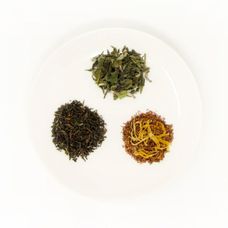 3 small piles of dry leaves of fine loose leaf teas sit on a white plate. These are examples of teas included in a tea subscription
