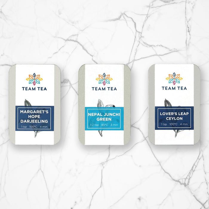 3 silver metal tins on a marble background. The tins have white paper labels with colourful banners on them and the names of teas