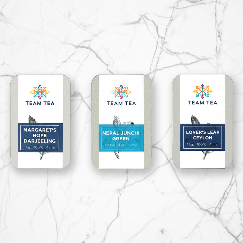 3 silver metal tins on a marble background. The tins have white paper labels with colourful banners on them and the names of teas
