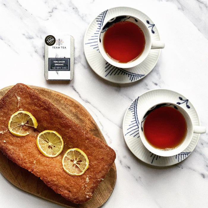 A homemade lemon drizzle cake sits next to 2 delicate tea cups of black tea. There is a metal tin of loose-leaf tea labelled Temi Sikkim Organic