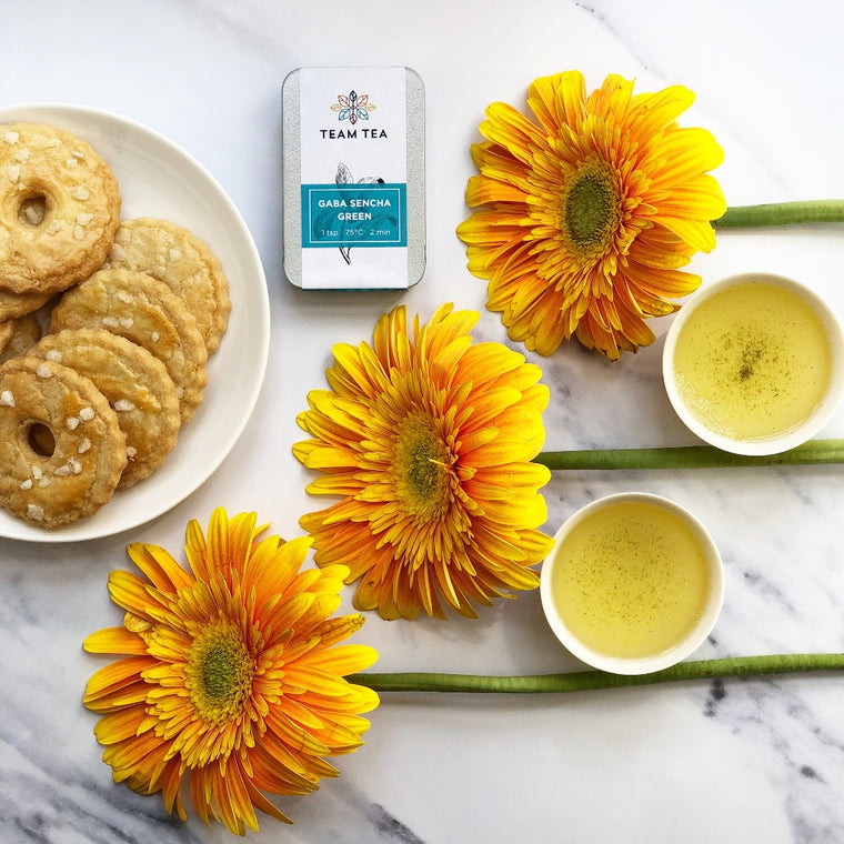 Three yellow sunflowers are arranged in the centre of the image next to 2 cups of green tea and a plat of round shortbread biscuits. There is a metal tin of loose leaf tea labelled GABA Sencha Green.