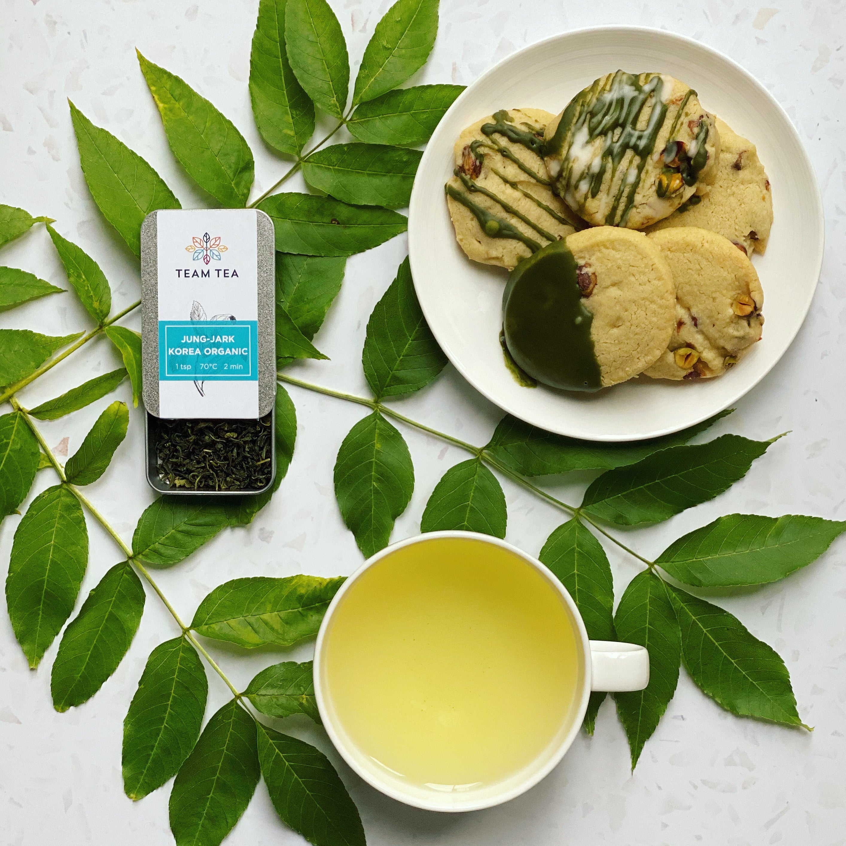 A cup of green tea with a tin of loose leaf green tea labeled 'Jung-Jark Korea Organic', served with matcha and pistachio cookies, presented on a plate surrounded by green tea leaves on a white background.