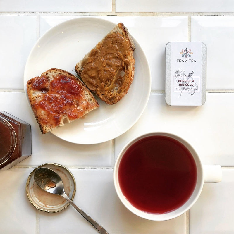 A cup of red herbal tea, next to a white plate with 2 small slices of toast, one with peanut butter, the other with jam. There is a metal tin containing loose-leaf Berries & Hibiscus herbal infusion