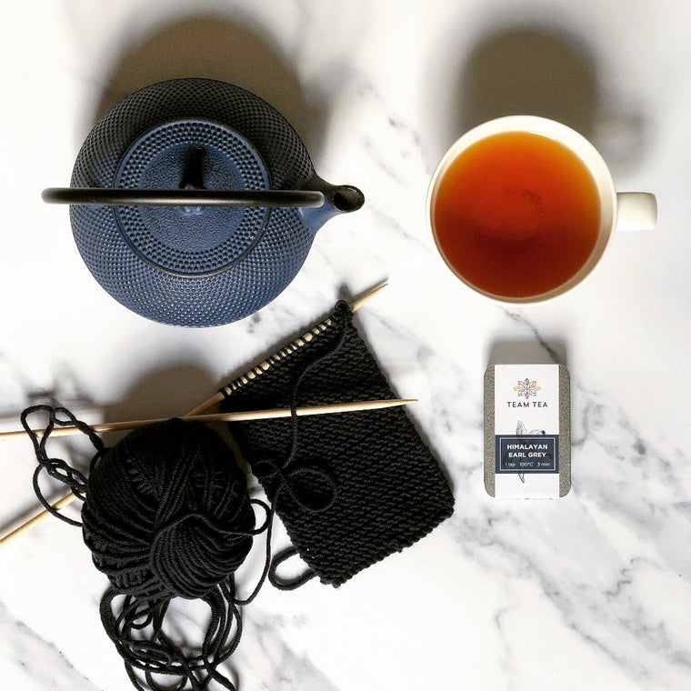 A brewed cup of black tea sitting next to a blue cast iron teapot and some work-in-progress knitting. There is a metal slide tin containing loose-leaf tea.
