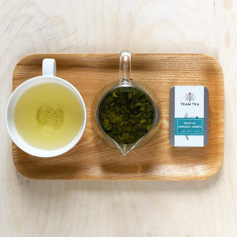 A rectangular wooden Japanese tea tray with a cup of brewed green tea, a small pitcher of loose-leaf Japanese sencha brewing and a tin of tea labelled Sencha Organic Green.
