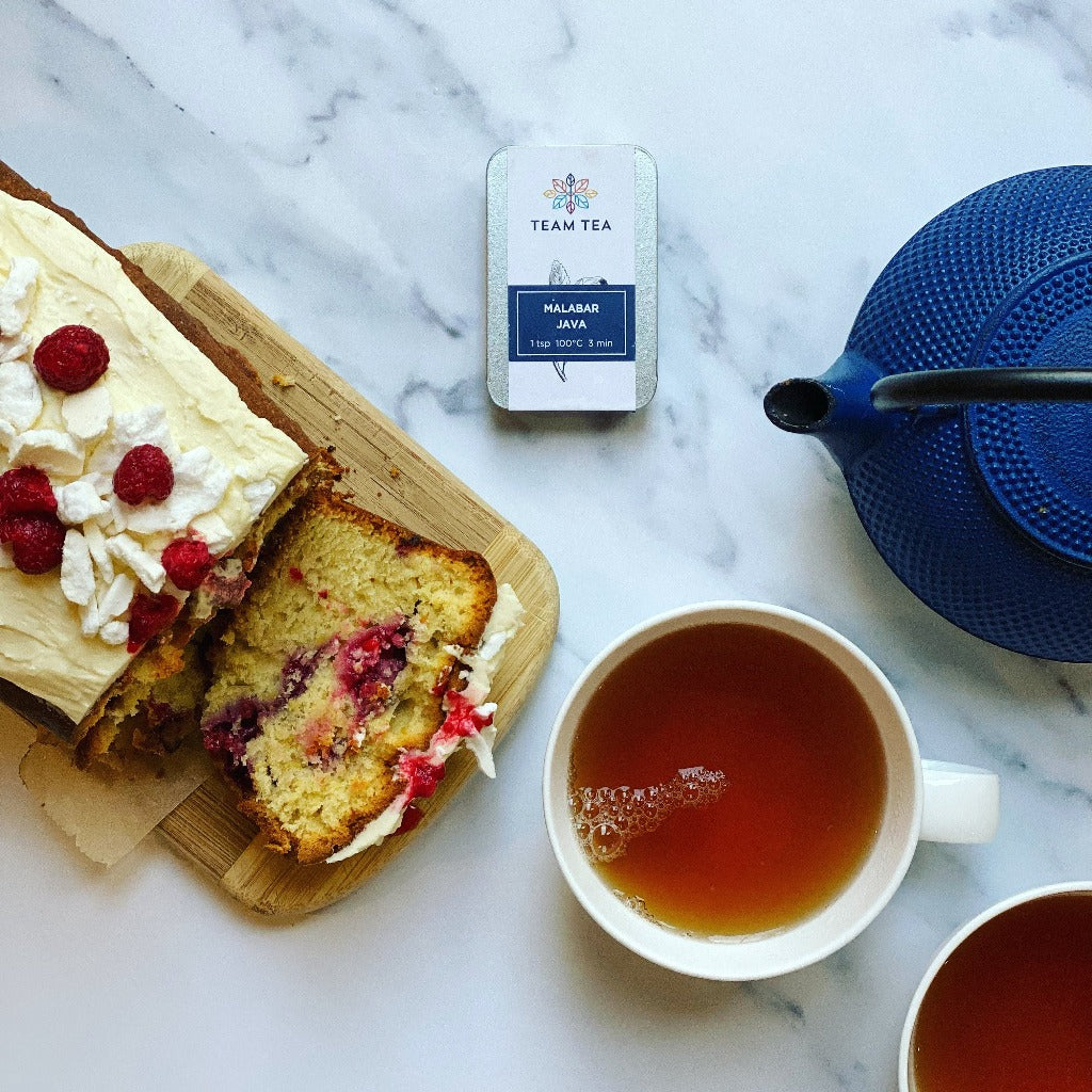 A cup of tea with a slice of cake on a wooden board, with a box of tea leaves labeled 'Malabar Java' in the background, and a blue cast iron teapot to the side.