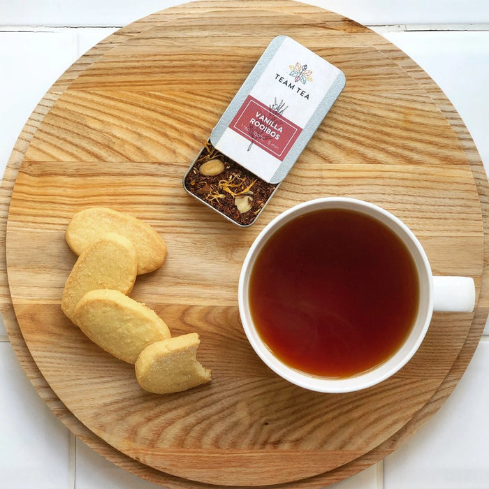 A cup of vibrant rooibos sits on a circular wooden tray with 4 shortbread biscuits and an open metal tin of loose-leaf vanilla rooibos tea.