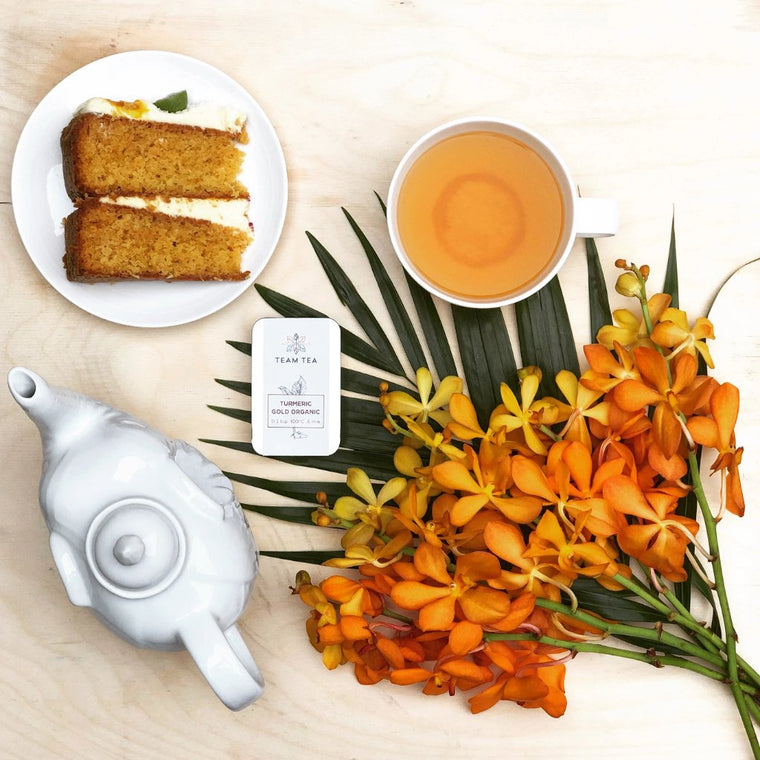 A cup of warm amber coloured herbal tea sits next to a slice of cake, and elephant shaped teapot, some striking orange flowers and a tin of loose leaf turmeric tea.