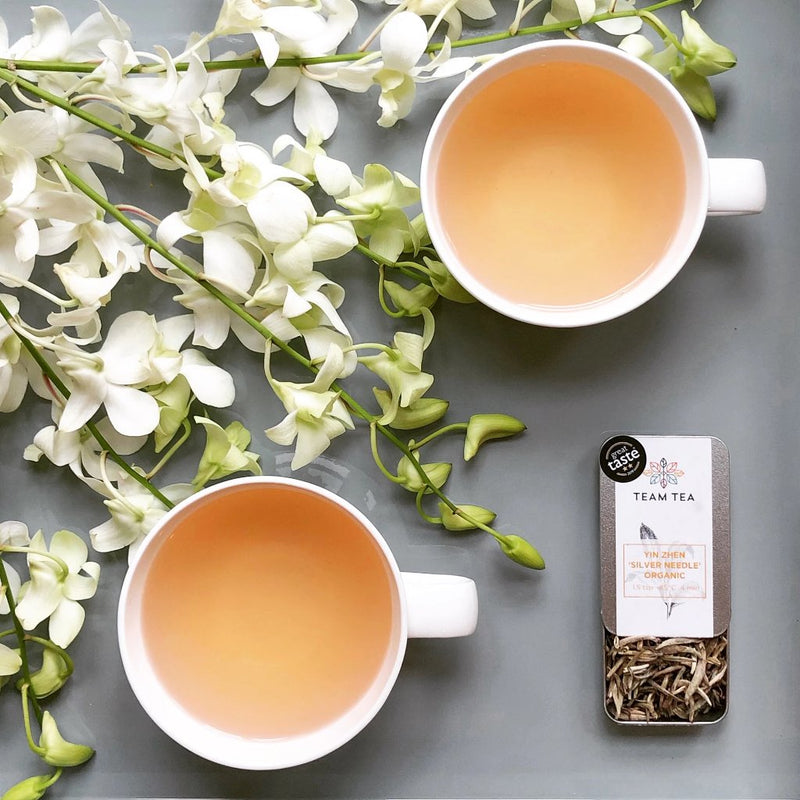 2 teacups of brewed white tea sit on a grey tray next to an open metal tin showing loose white tea buds. There are white flowers arranged on the tray.