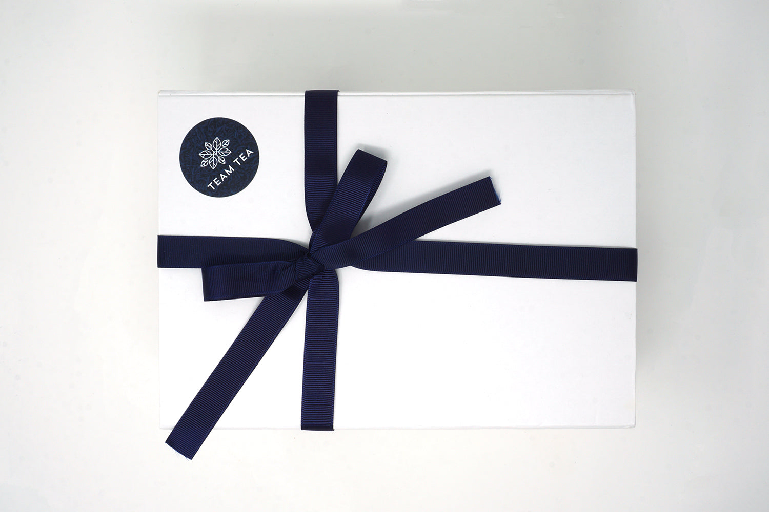 A white rectangular gift box with a navy blue ribbon, tied in a bow on the front, containing tea slide tins.