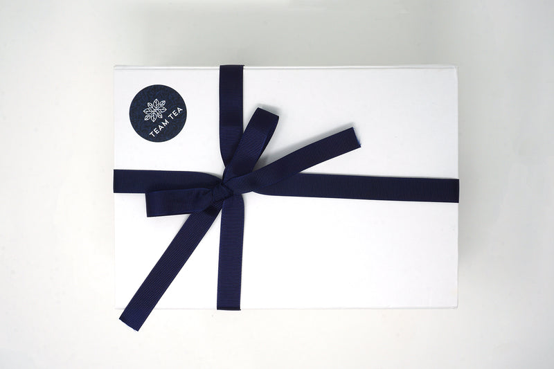 A white rectangular gift box with a navy blue ribbon, tied in a bow on the front, containing tea slide tins.