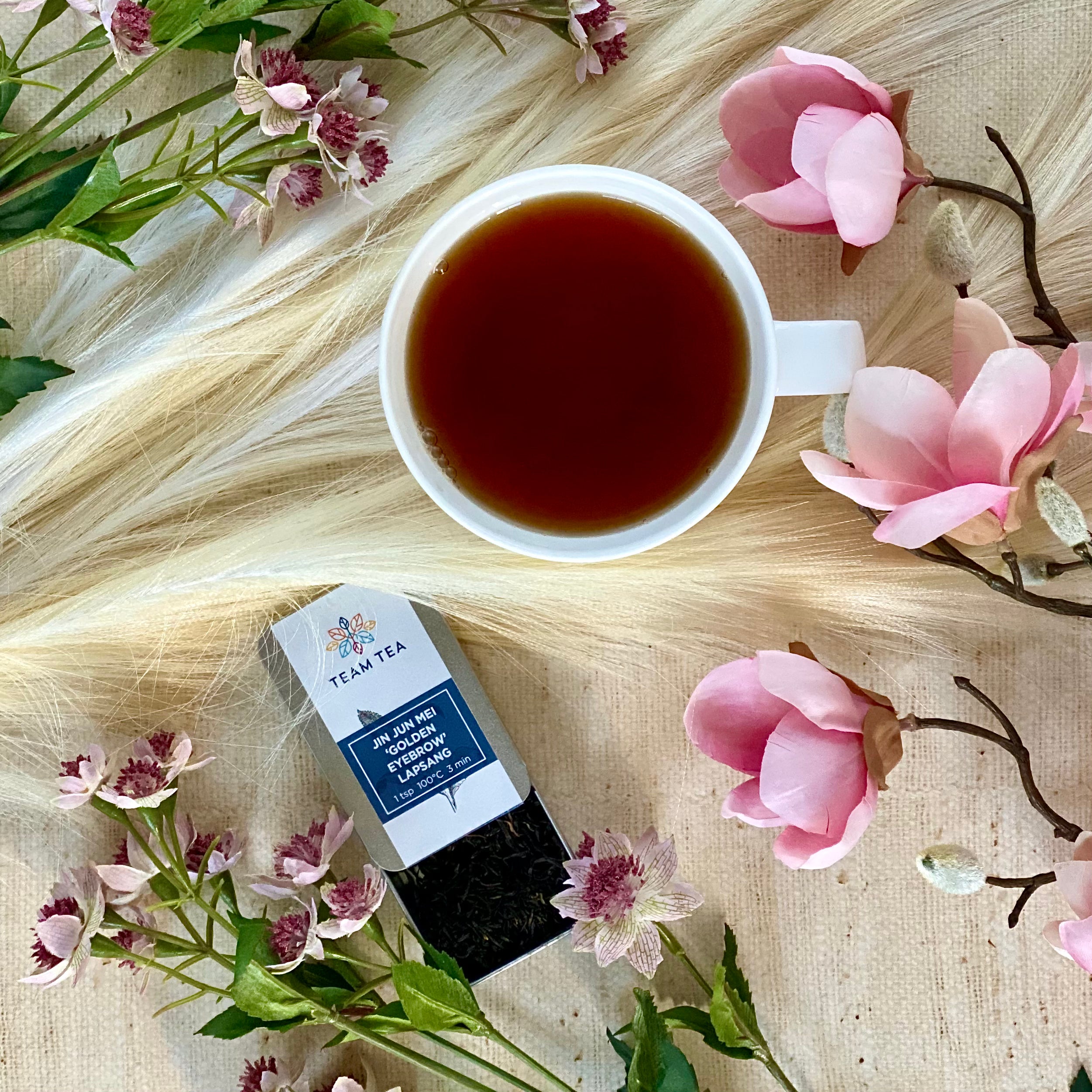 Pink flowers laid on a linen background with cup of black tea and tin of Jin Jun Mei Golden Eyebrow Lapsang