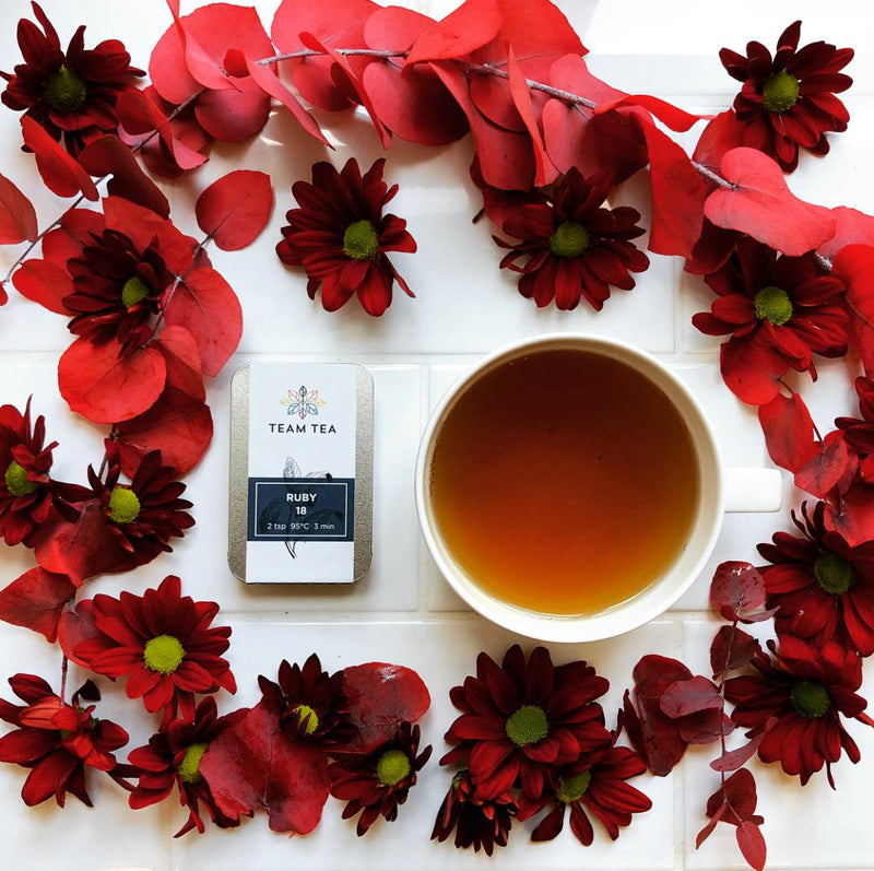 A cup of black tea sits on a white tiled background surrounded by a ring of red flowers. There is a metal tin of loose-leaf tea from Taiwan.