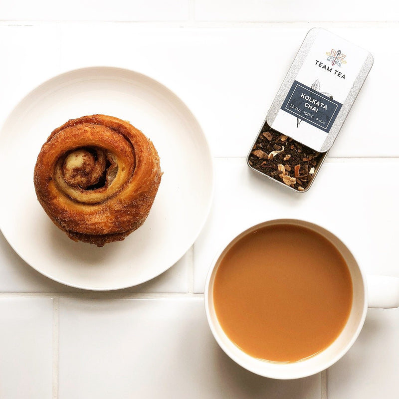 A mug of milky chai next to a white plate with a cinnamon bun and in the top right of the image is an open tin showing the loose-leaf spiced chai blend.