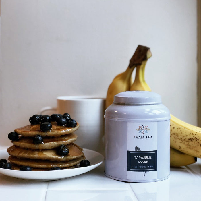 A stack of fluffy pancakes drizzled in maple syrup and fresh blueberries next to a caddy of loose-leaf tea labelled Tarajulie Assam. There is a mug of black tea and a bunch of bananas in the background.