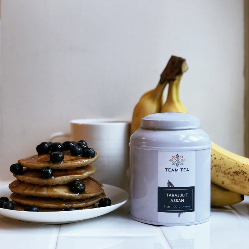 A stack of fluffy pancakes drizzled in maple syrup and fresh blueberries next to a caddy of loose-leaf tea labelled Tarajulie Assam. There is a mug of black tea and a bunch of bananas in the background.