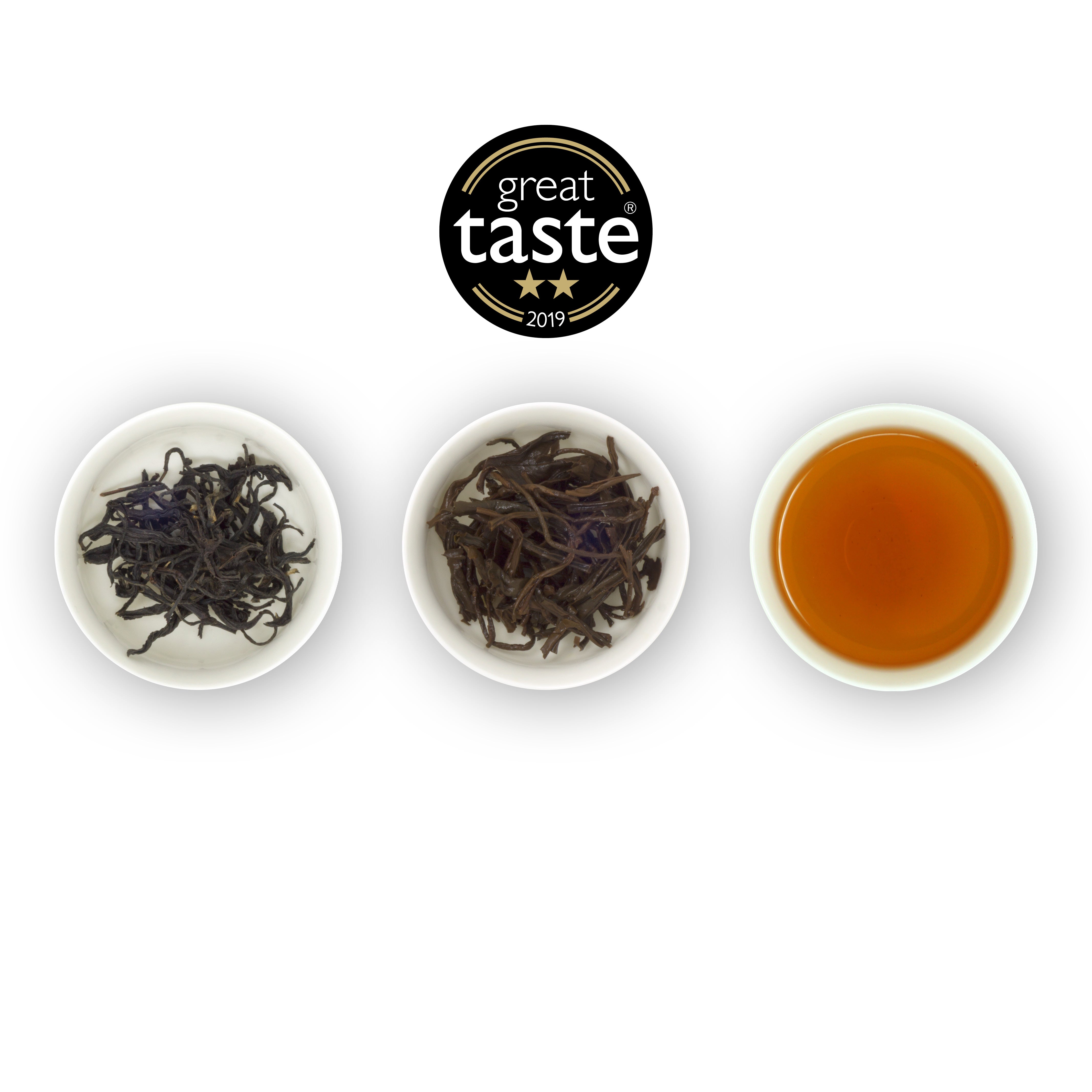 Three circular bowls arranged side by side with the left bowl containing dry tea leaves, the middle bowl showing wet tea leaves, and the right bowl displaying a cup of brewed black tea. Above is the Great Taste Awards logo