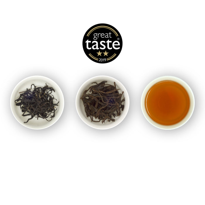 Three circular bowls arranged side by side with the left bowl containing dry tea leaves, the middle bowl showing wet tea leaves, and the right bowl displaying a cup of brewed black tea. Above is the Great Taste Awards logo