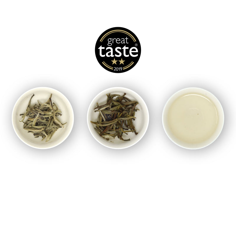 Three circular bowls with samples of Yin Zhen Silver Needle tea. The first bowl shows dried silver needle buds, the second bowl shows the tea leaves after brewing, and the third bowl shows the brewed tea liquid. Above is the Great Taste Awards logo