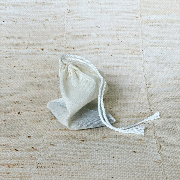 A beige unbleached cotton teabag with a drawstring, placed on a textured background.