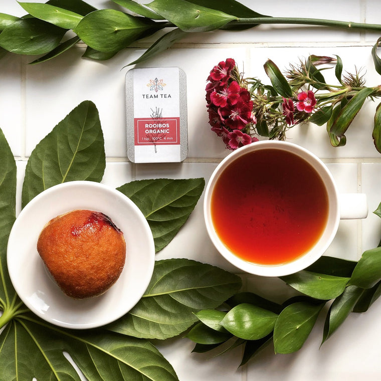 A cup of brewed vibrant red rooibos next to a doughnut on a small white plate and a metal tin of loose-leaf tea. Surrounded by verdant green foliage