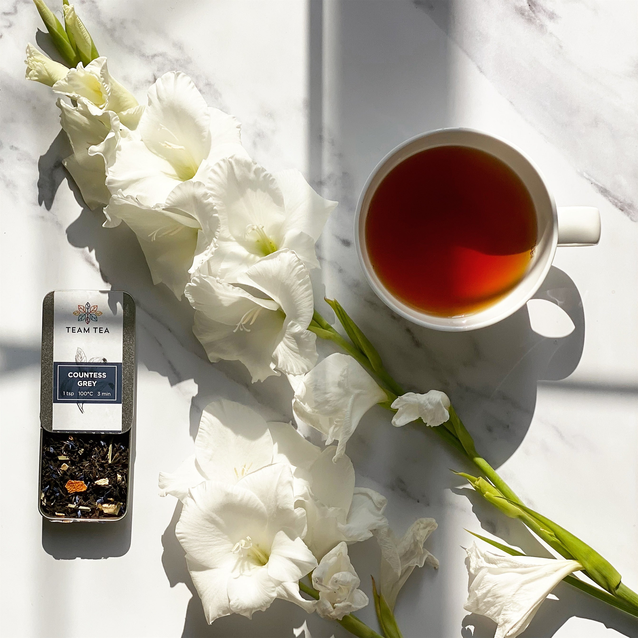 A cup of black tea placed next to an open tin of loose leaf tea and flowers on the side, placed on a marble surface.