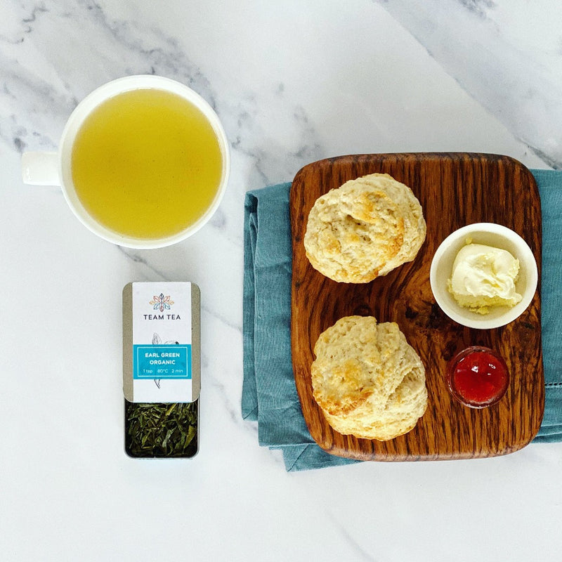 open tin of Earl Green Organic, showing the tea leaves. Alongside the tin is a cup of brewed green tea and a wooden chopping board with 2 scones, a small ramekin of clotted cream and some jam 