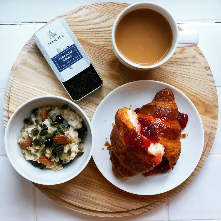 A styled breakfast scene on a round wooden tray with a cup of tea, a croissant with jam, an oatmeal bowl with nuts and seeds, and a tin of Team Tea Tarajulie Assam.