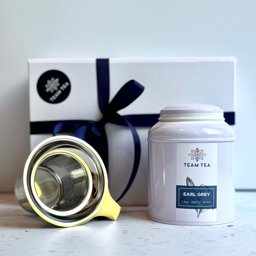 A caddy (regular tin) of Team Tea Earl Grey next to a strainer basket in front of a gift box and white background