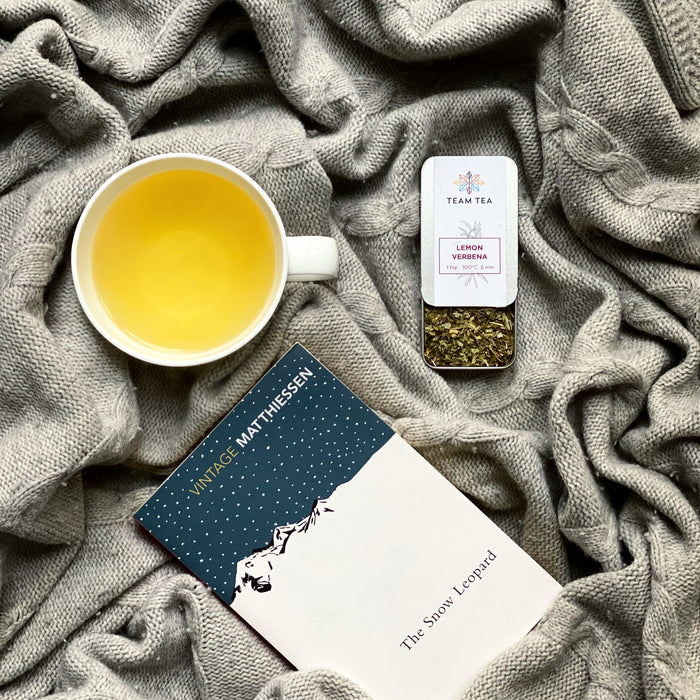 A cup of lemon verbena tea is placed on a knitted grey blanket next to a tin of loose tea leaves, with a book titled 'The Snow Leopard'