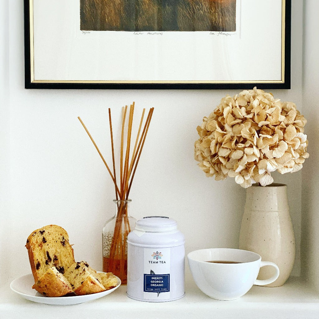 Caddy of Imereti Georgia Organic with a cup of tea and a chocolate panettone sitting on a shelf with flowers, a reed diffuser and a framed print on the wall